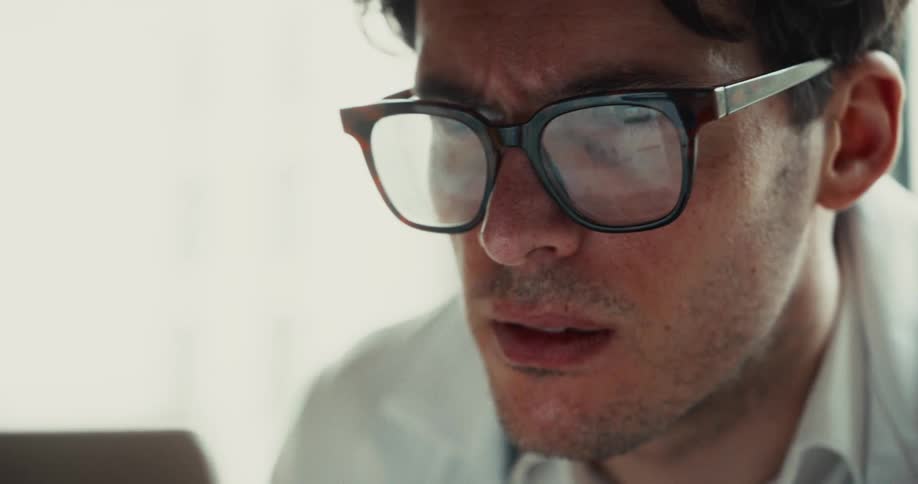 A young man wearing a white shirt and thick glasses looks intently at a screen with a focused and slightly stressed expression in a bright indoor environment