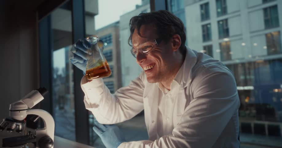 In this video a scientist in a lab coat and safety glasses laughs while holding up a flask containing an ambercolored liquid he is seated in a laboratory next to a microscope looking at the flask with an expression of triumph or excitement