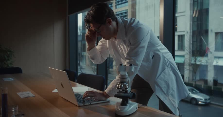 A scientist in a white lab coat works intently at a laptop late in the evening by a large windowbeside the computer sits a microscope highlighting a blend of digital data analysis and traditional laboratory research