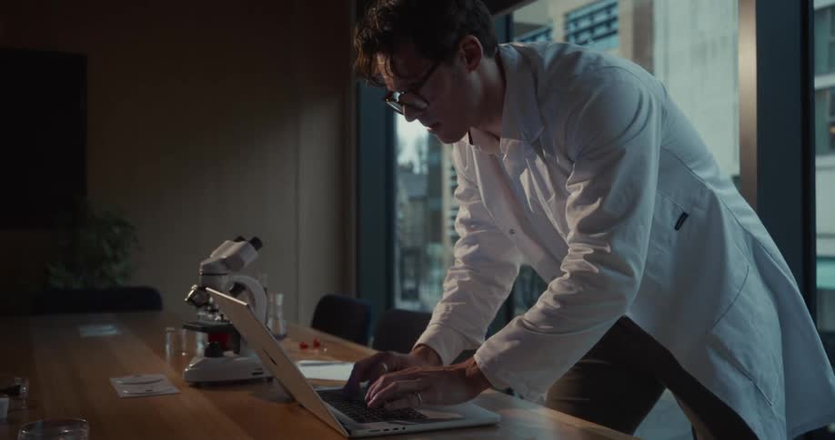 A man in a lab coat wearing eyeglasses is standing typing urgently on a laptop in a dimly lit room