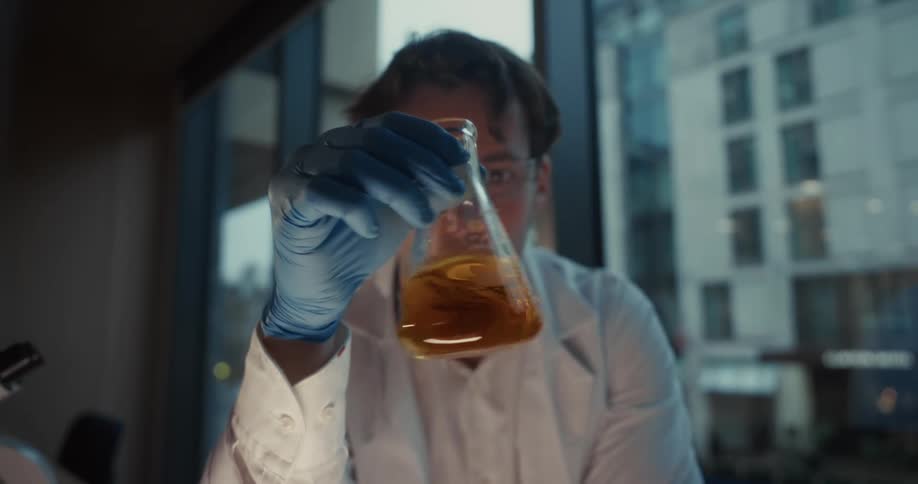Researcher in a lab coat and blue gloves holds a flask with orange liquid in front of a window with buildings in the background