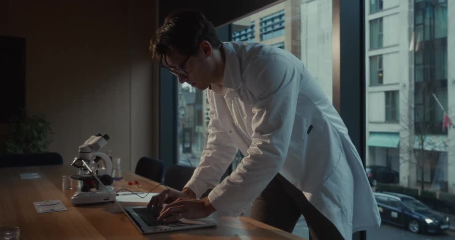A rushed man in a white lab coat is typing on a laptop compute in a room with a long desk and a microscope