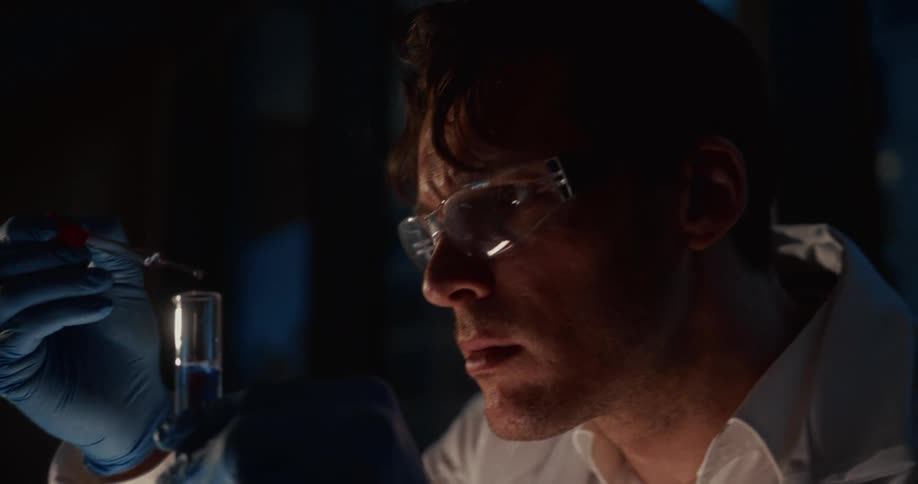A man wearing googles gloves and a lab coat is holding a test tube and dropper dropping liquid into the test tube in a dark room