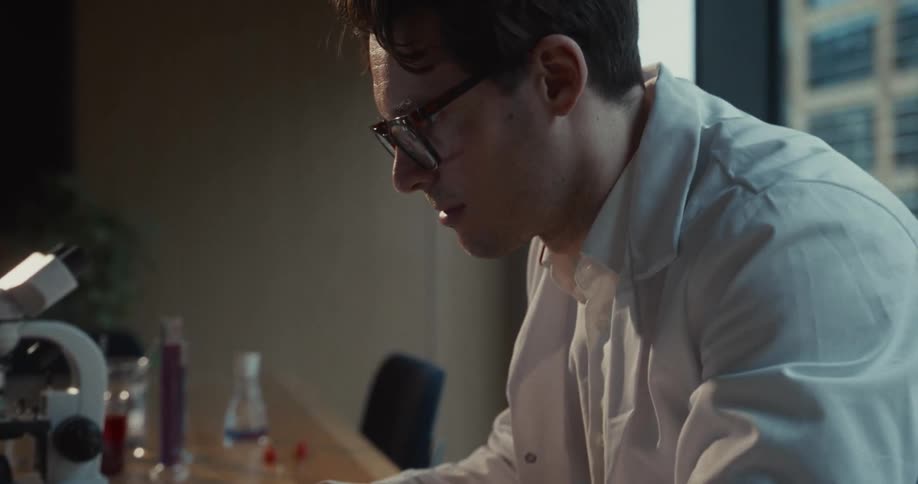 Researcher in a lab coat types on a laptop at a table with a microscope and flasks in the background