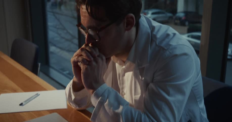 In the video a man wearing a white lab coat and glasses sits at a wooden desk in front of a laptop he transitions from typing on the keyboard to resting his chin on his clasped hands appearing deep in thought or concerned