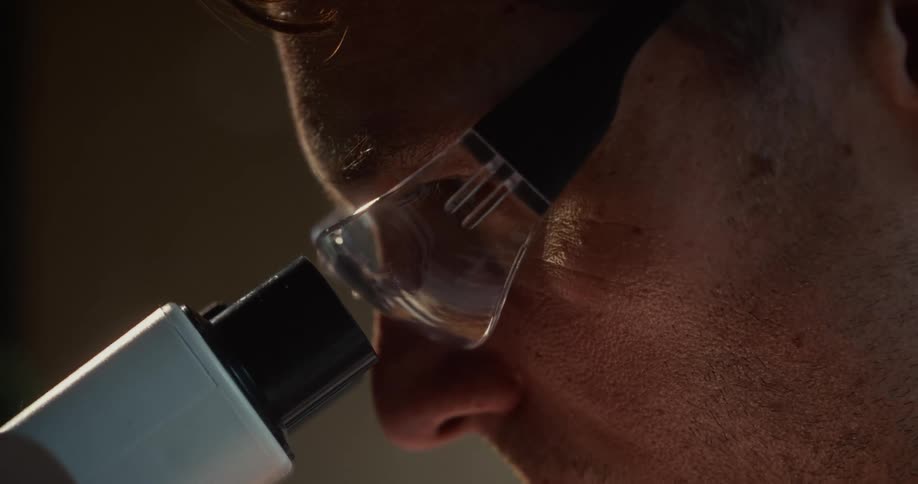 Man wearing safety goggles observes a sample through a microscope in a laboratory setting