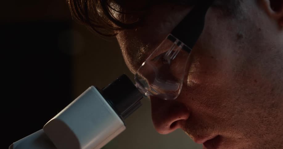 A close view shows a scientist wearing clear safety glasses looking intently through the eyepiece of a white microscope in a dark laboratory