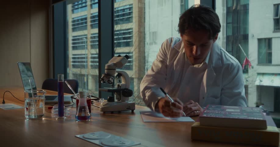 A male scientist in a white lab coat examines a sample through a microscope and writes notes in a modern laboratory