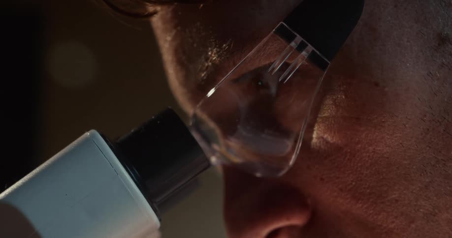 A close view shows a scientist wearing clear safety glasses as he looks intently through the eyepiece of a white microscope in a dark laboratory setting