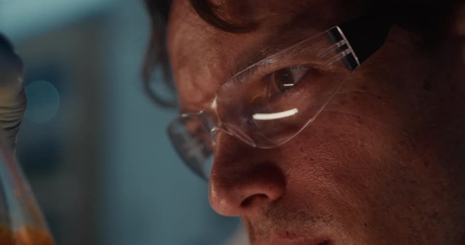A close view shows a scientist wearing clear safety glasses and a white coat looking intently at a glass flask containing amber liquid in a laboratory