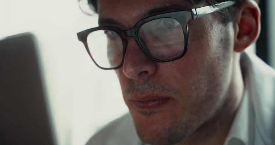 A close view shows a doctor wearing black rimmed glasses and a white coat looking at a screen with a serious expression in a bright room