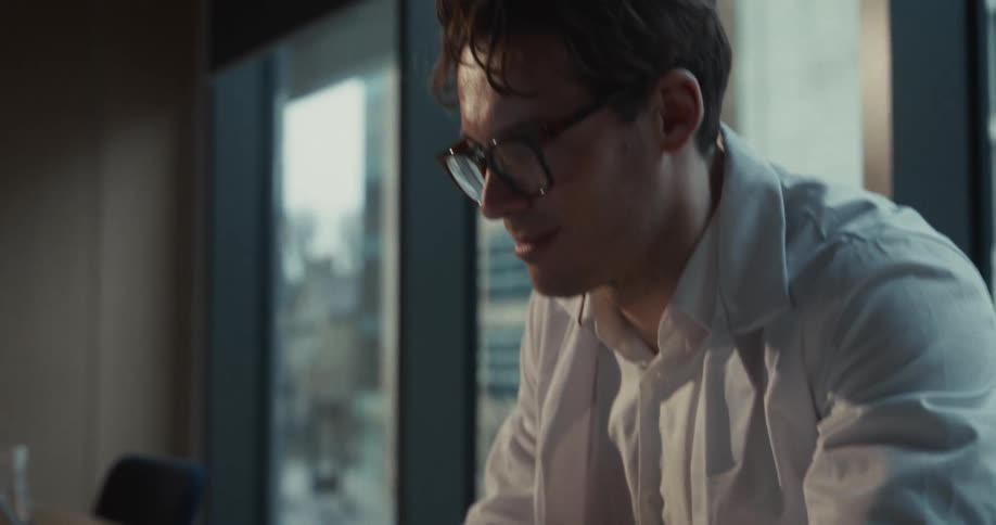 A man wearing a white lab coat types on a laptop computer in a dimly lit office