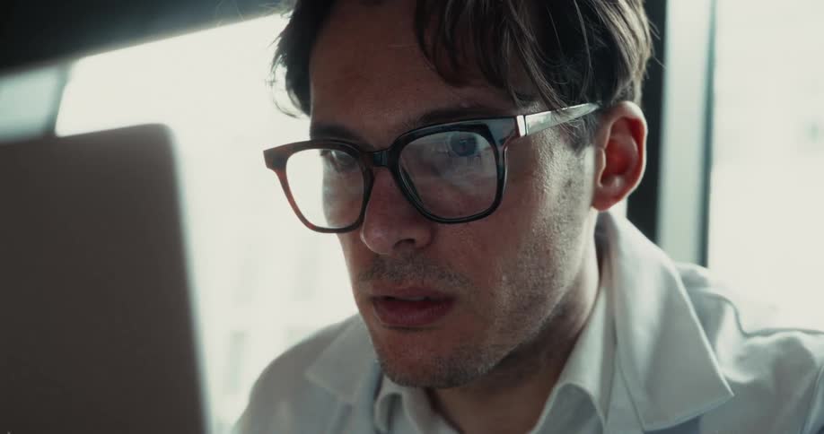 A man in a white lab coat and glasses looks at a laptop screen