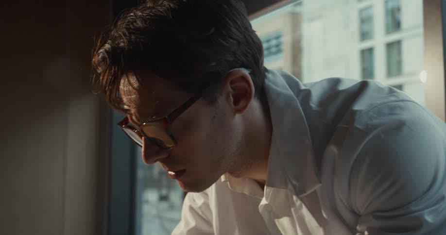 A male scientist in a white lab coat puts on his glasses and leans over a workspace in a laboratory