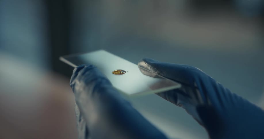 A person wearing blue nitrile gloves holds a glass slide while a pipette dispenses a single drop of amber colored liquid