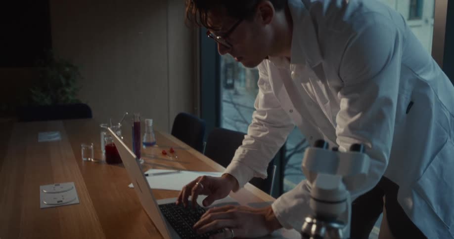 Scientific technician in a conference room with a microscope and some chemicals works on a laptop