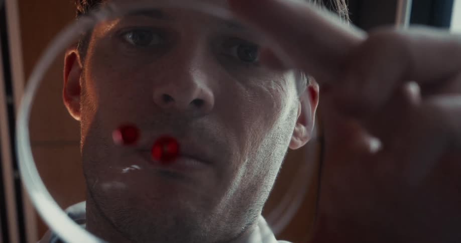 An up close view of a man concentrating on using a pipette to drop red droplets on a clear petri dish near his face