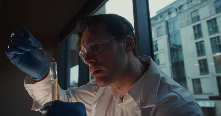 Researcher wearing gloves and safety goggles uses a pipette to handle liquid in a test tube near a window