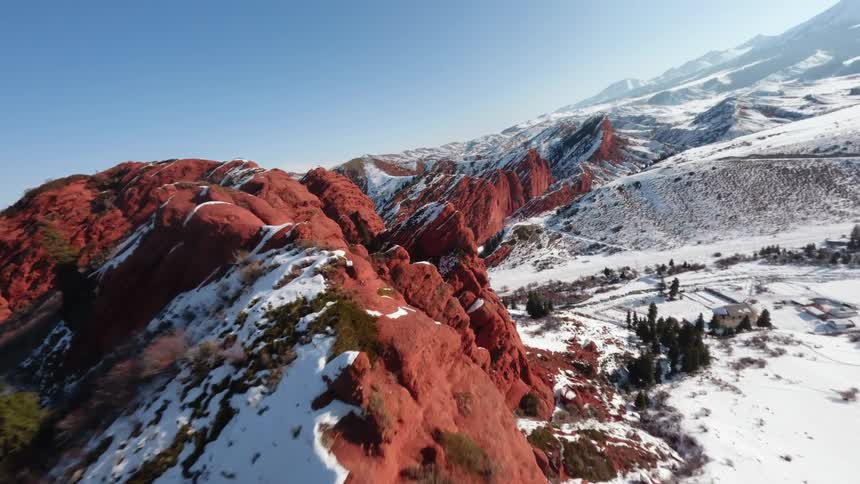 An fpv drone captures a highspeed flight over striking red rock formations dusted with snow under a clear blue skythe camera weaves through narrow gaps in the jagged cliffs revealing a vast wintry valley and distant mountain ranges