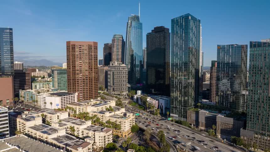 A high view shows many tall glass skyscrapers and office buildings in Downtown Los Angeles under a clear blue sky as many cars move along a wide highway during the day