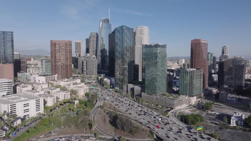 An aerial view of Los Angeles skyscrapers and office buildings under a clear blue sky as many cars move along a wide highway during the day