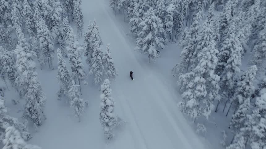 An aerial shot follows a person riding a bicycle on a snowy trail through a forest of snow covered trees toward a lit cabin
