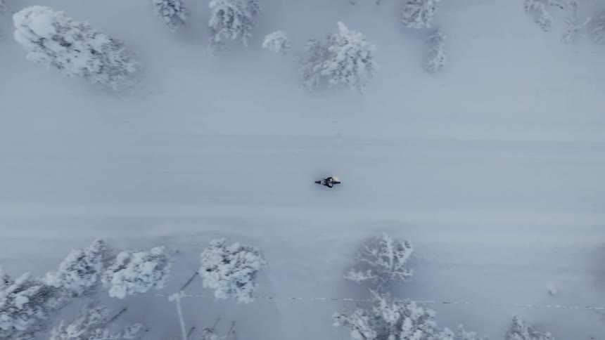 A person walks slowly down the center of a narrow snowcovered road flanked by dense frostladen trees the topdown aerial perspective captures the stark contrast between the lone figure and the vast white winter landscape