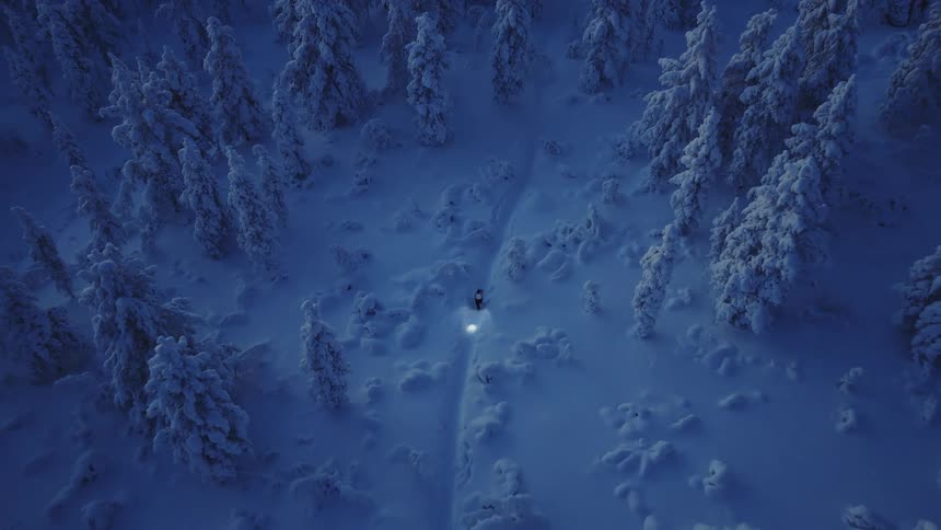 An aerial shot captures a lone figure with a bright headlight navigating a narrow trail through a dense snowcovered forest at twilightthe deep blue hues of the landscape contrast with the warm glow of the light as it illuminates the thic