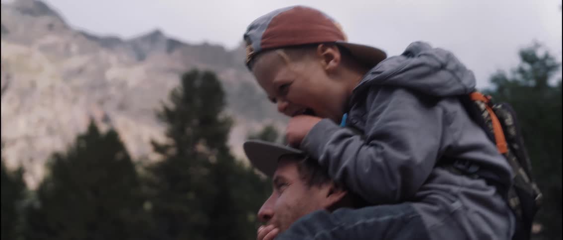 A boy sits on a mans shoulders and covers the mans mouth playing while they walk outdoors near a forest and mountains