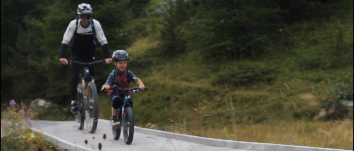 A boy rides his bicycle along a wooden path while an adult cycles behind him and a dog runs ahead on the trail