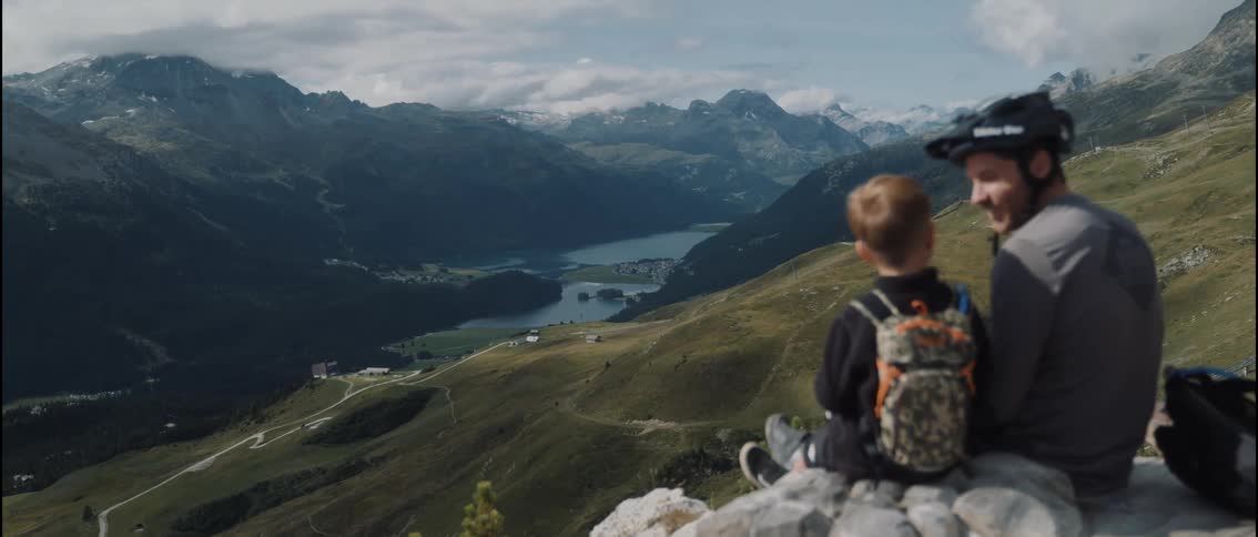 A father and son sit together on a mountainside overlooking a valley