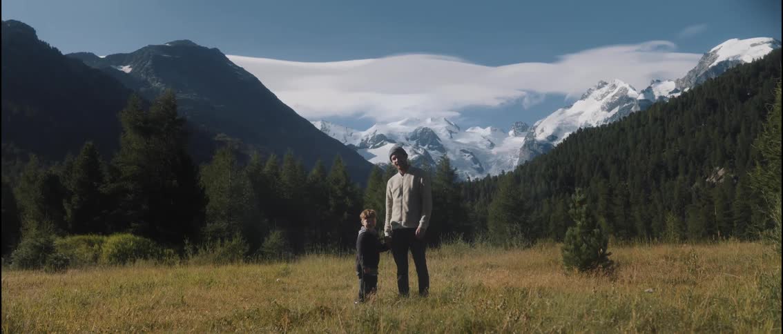 A man and a young boy stand together in a grassy field holding hands while facing the camera with a massive snow capped mountain range and dense evergreen forest under a bright sky in the background