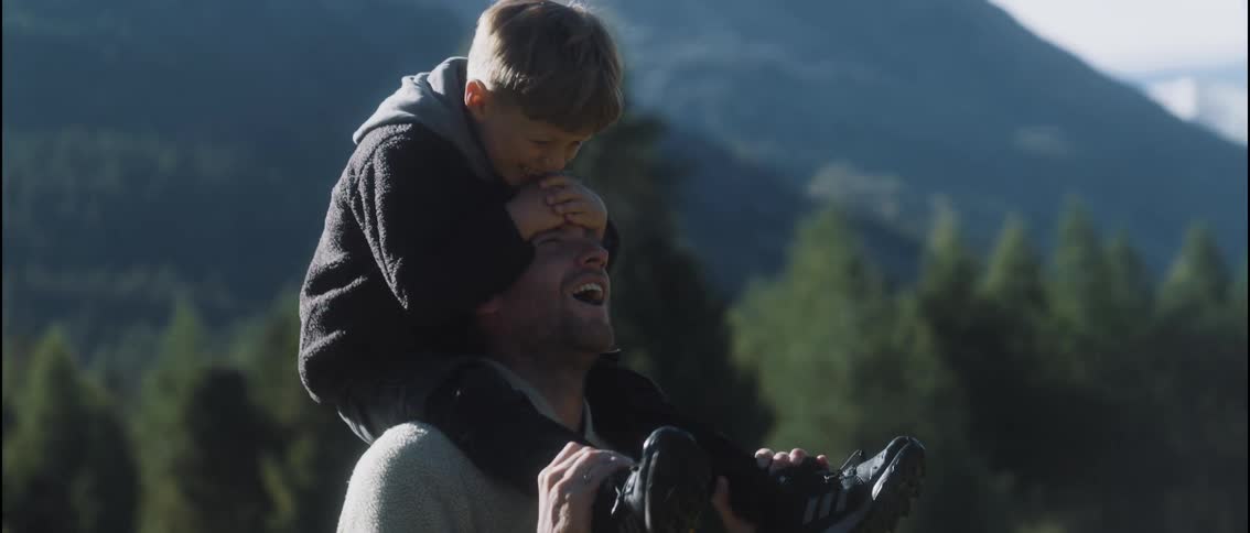 A father carries his son on his shoulders in a grassy field outdoors as they both laugh and enjoy the moment together