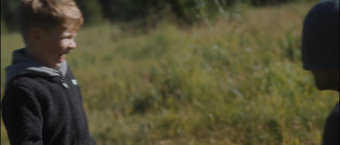 A smiling young child playfully leans over and pulls the beanie over a mans face in a sunny grassy field