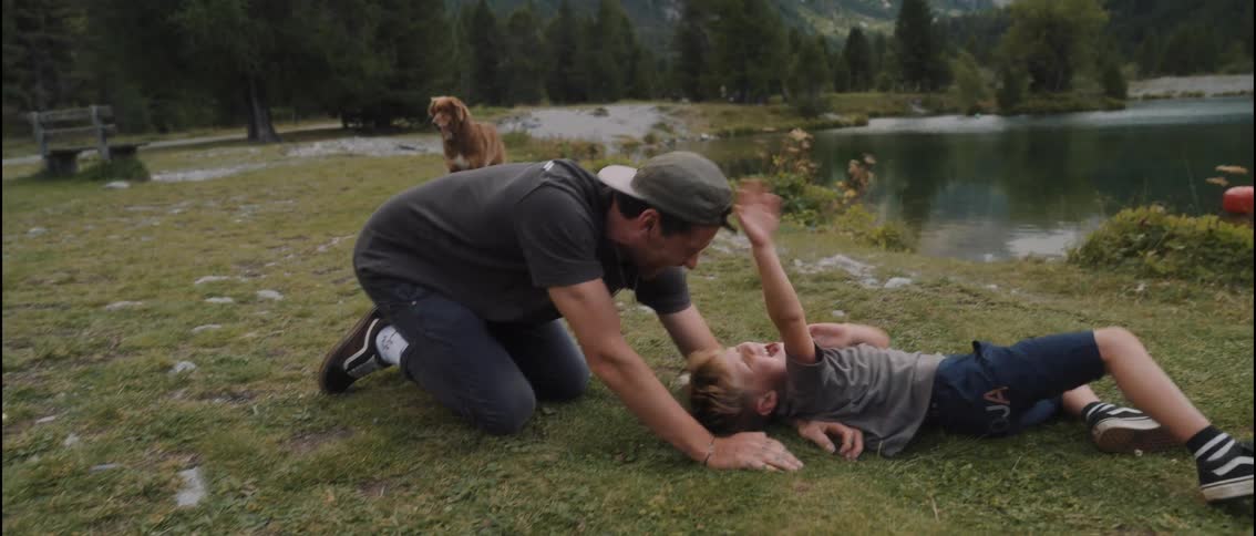 A man in a baseball cap playfully tickles a young boy on a grassy bank beside a mountain lake while a dog stands in the background near a wooden bench and evergreen trees