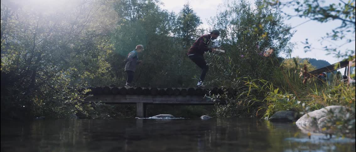 An adult carrying items and a child leap on a small wooden bridge over a calm sunlit river surrounded by lush greenery