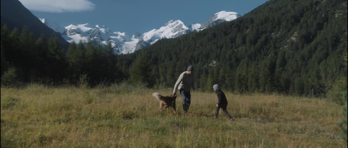 Man plays with his young son and their dog on a grass covered mountain surrounded by forested mountains
