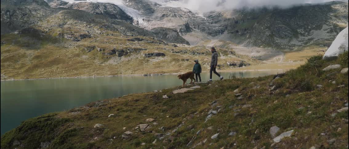 A man and a child walk with a dog along a grassy ridge overlooking a mountain lake
