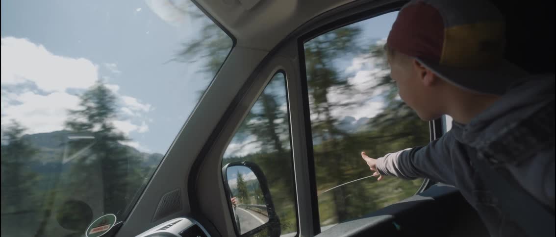 A boy puts his hand and arm out of the car window as the vehicle drives through a forested mountain road