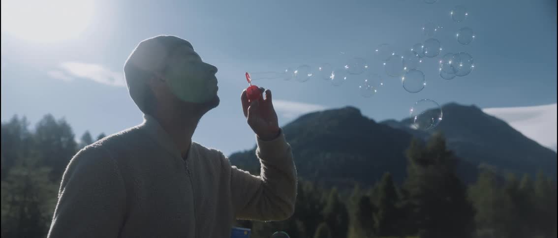 A silhouetted man in a beanie gently blows soap bubbles into the sunlit mountain air with forested peaks and a bright blue sky in the background