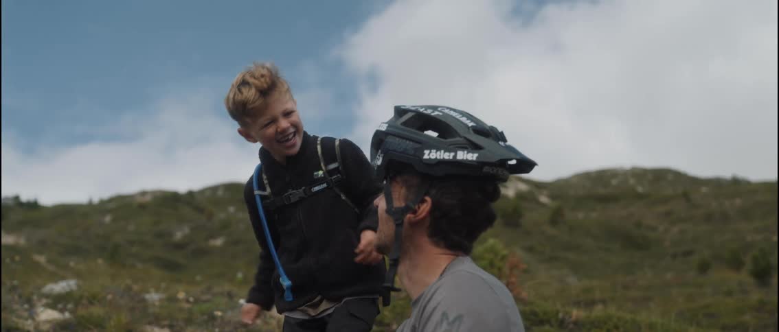 A young boy playfully adjusts a mountain bike helmet on a mans head while they sit together on a grassy hillside the child laughs claps his hands and reaches out toward the mans face in a joyful candid moment outdoors