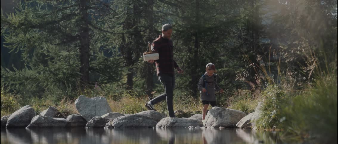 A man and a child walk across large rocks along the shore of a pond in a forest