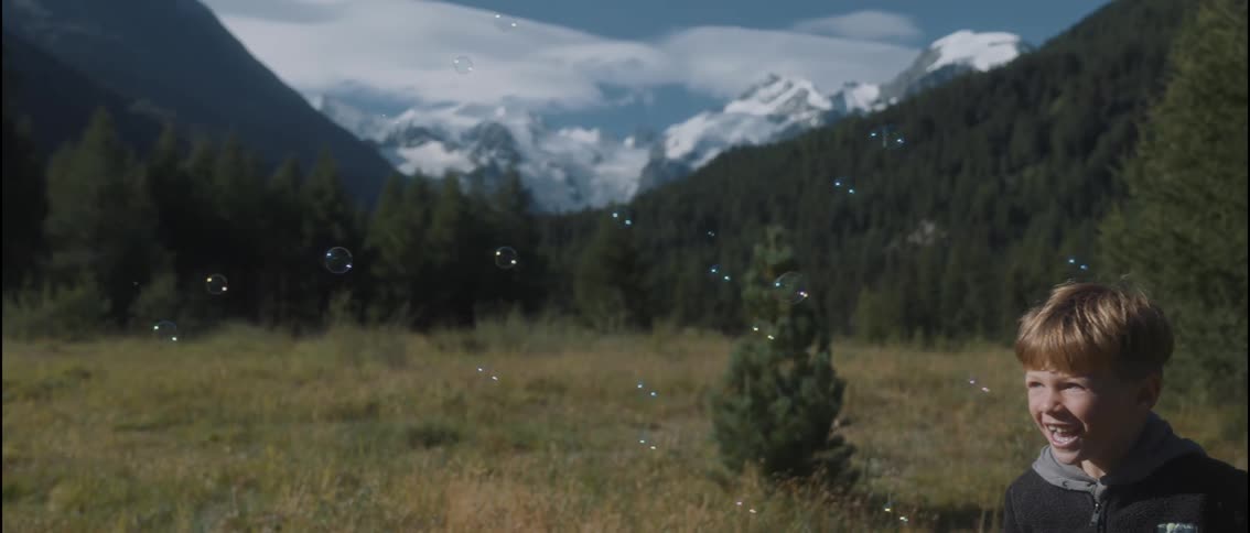 A young boy in a dark fleece hoodie chases and pops floating bubbles in a sunny mountain meadow he smiles and laughs with excitement while snowcapped peaks and a dense evergreen forest provide a scenic backdrop