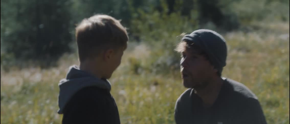 A young boy in a black hoodie playfully pulls a grey beanie over a smiling mans eyes while they sit together in a sunlit grassy field during the daytime