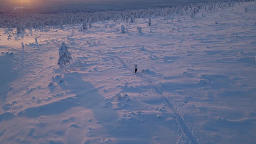 A lone figure walks across a vast snowcovered landscape at sunset leaving a trail of footprints among scattered frostcovered trees