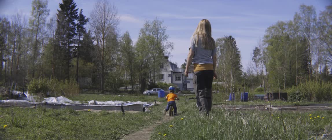 A woman claps and cheers as a young toddler boy rides his bicycle outdoors.