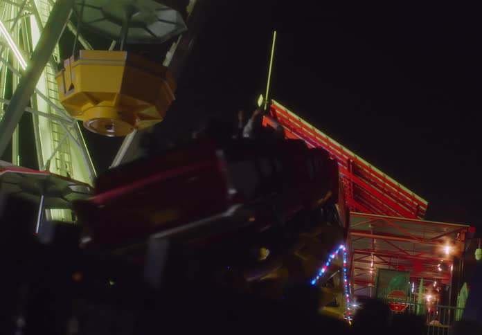 A spinning carnival ride moves quickly in the foreground while a ferris wheel stands tall under the night sky at a crowded amusement park filled with glowing colored lights and metal structures