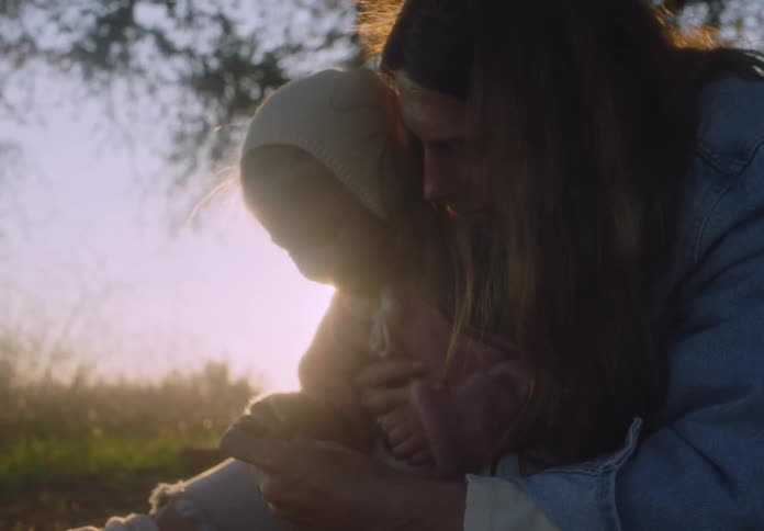 A woman with long dark hair wearing a denim jacket sits in a field and holds a small child wearing a tan bonnet as golden sunlight filters through the trees during a calm afternoon
