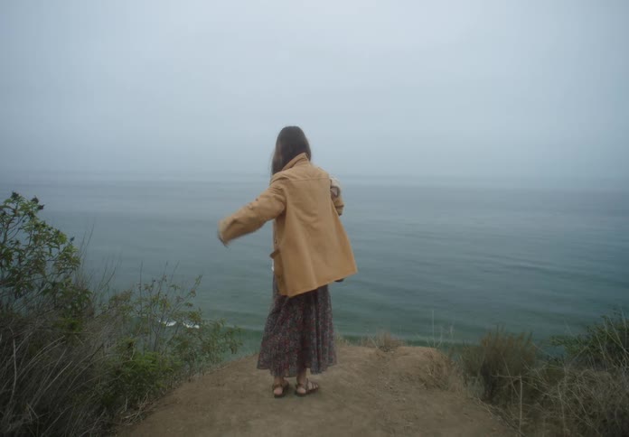 A woman stands on a dirt cliff overlooking a foggy ocean while holding a small child who wears a beige coat