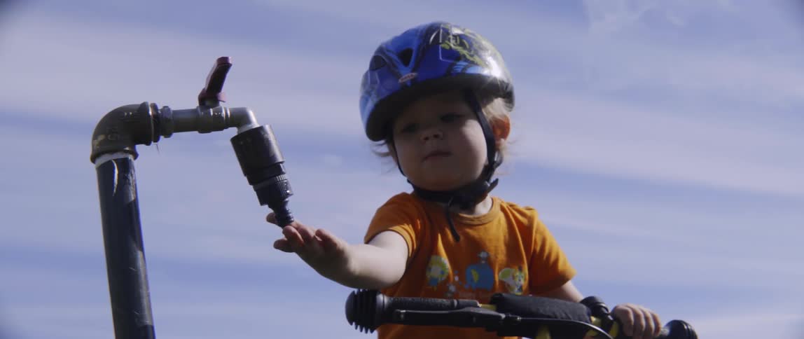A toddler boy is wearing a bicycle helmet and reaching toward a spigot